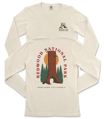 a cream-colored long-sleeved t-shirt with a graphic design depicting a tall tree trunk and a sunset. The design includes the text "REDWOOD NATIONAL PARK" and "NORTHERN CALIFORNIA" printed on the back of the shirt.