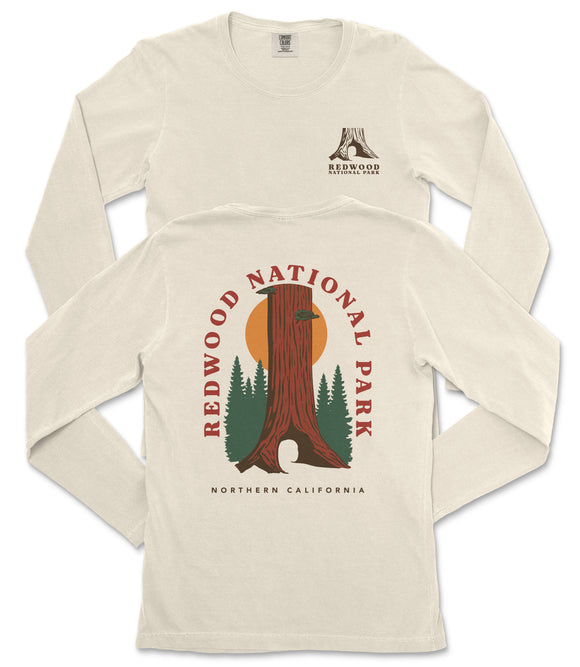 a cream-colored long-sleeved t-shirt with a graphic design depicting a tall tree trunk and a sunset. The design includes the text "REDWOOD NATIONAL PARK" and "NORTHERN CALIFORNIA" printed on the back of the shirt.