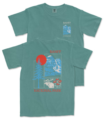 A green t-shirt with a graphic design featuring a mountain landscape, a red sun, and various wildlife elements. The text on the t-shirt reads "BANFF NATIONAL PARK".