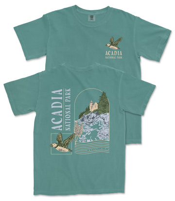 A green t-shirt with a graphic design depicting a bird and a landscape with a building, along with text that says "ACADIA NATIONAL PARK".