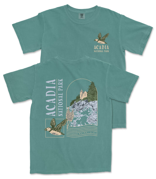 A green t-shirt with a graphic design depicting a bird and a landscape with a building, along with text that says "ACADIA NATIONAL PARK".