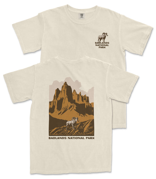 a white t-shirt with a graphic design depicting a mountain landscape with a goat in the foreground. The text on the t-shirt reads "Badlands National Park".