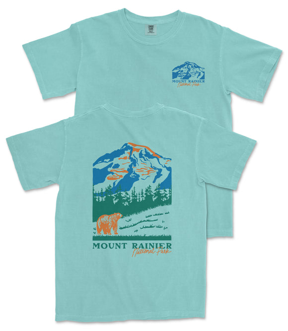 A light blue t-shirt with a graphic design depicting a mountain landscape, including a mountain peak, trees, and a bear. The text "MOUNT RAINER" and "National Forest" is printed on the t-shirt.
